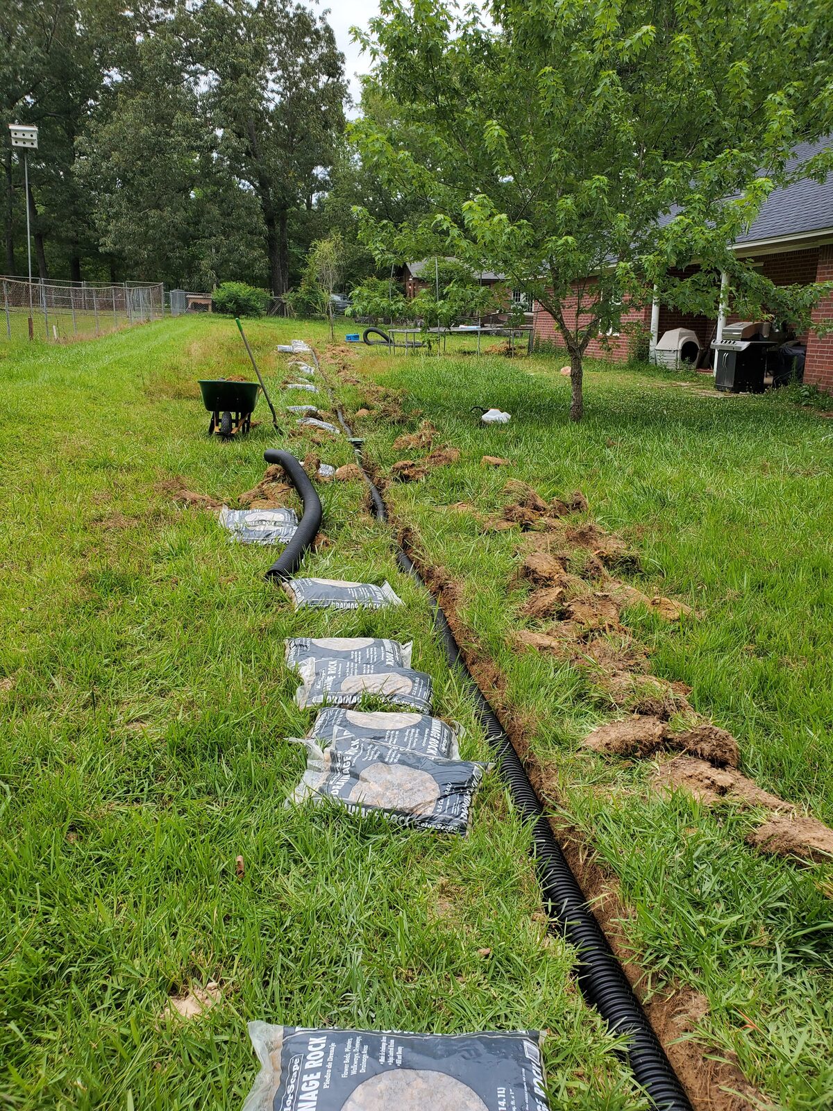 French drain installation in progress — open trench with perforated pipe and bags of drainage rock staged along the run